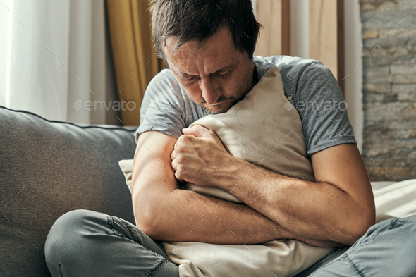 Depressed man sitting on living room sofa and hugging pillow