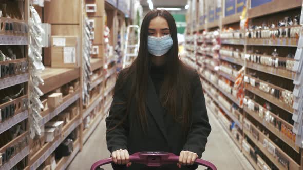 Woman wearing protection facemask choosing wallpaper in hardware store. Girl pushing shopping cart b
