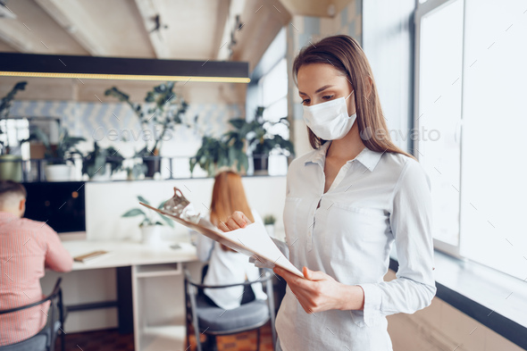 Young businesswoman in face mask standing in office and holding clipboard with documents