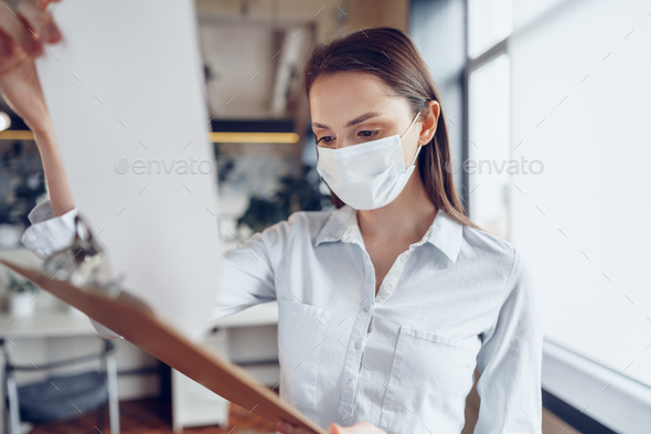 Young businesswoman in face mask standing in office and holding clipboard with documents
