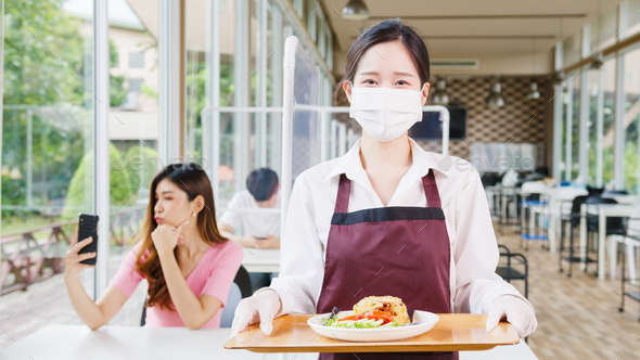 Female restaurant staff wearing protective face mask holding food tray to serving meal to customer.