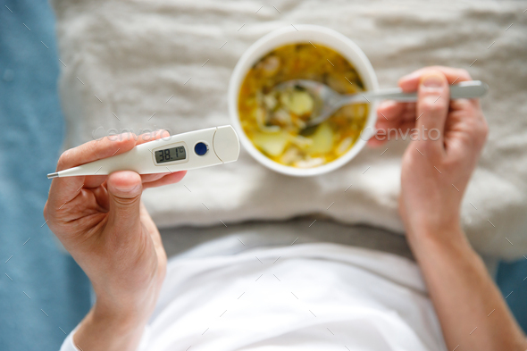 male eating healthy chicken soup to fight the flu sickness, holding thermometer