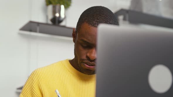 Young african man student taking notes in notebook using laptop alt