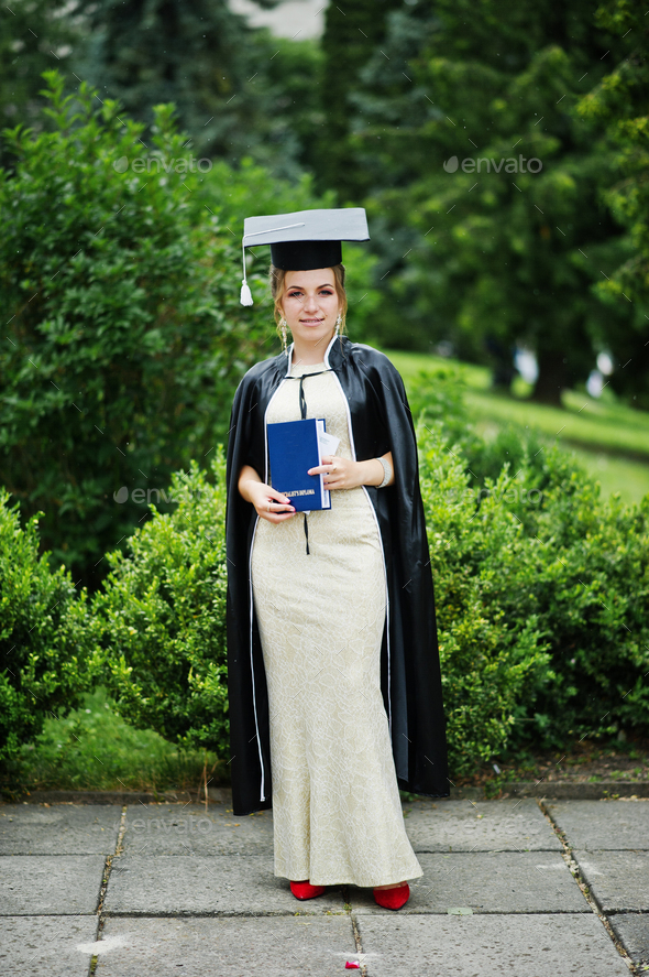Portrait of a beautiful female graduate in dress and graduation gown with hat posing in the park.