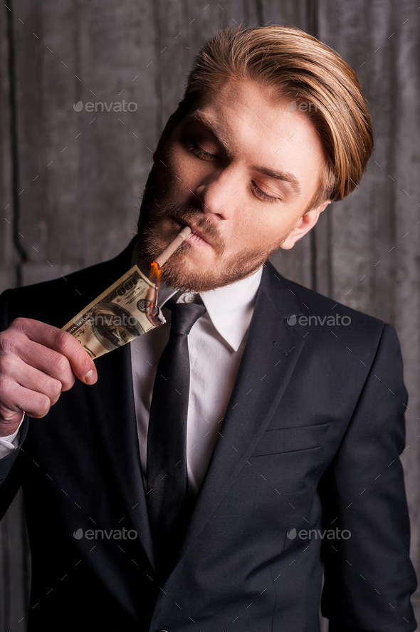Burning money. Handsome young man in formalwear lighting a cigarette using paper currency