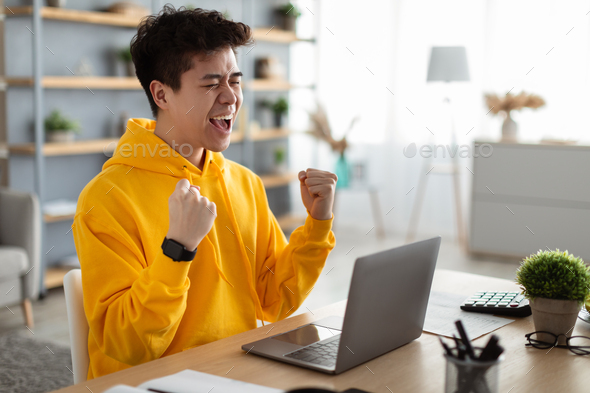 Asian guy using laptop celebrating success shaking fists