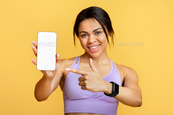 Sports application. Excited black lady holding and showing smartphone, indicating at black empty
