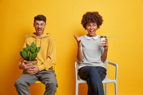 Annoyed mixed race woman and man scream loudly pose next to each other on comfortable chairs drink c