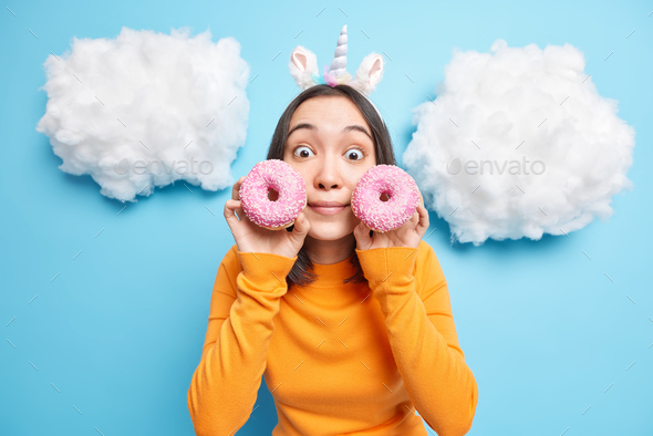 Photo of shocked brunette Asian girl holds two glazed donuts near face has bugged eyes surprised to