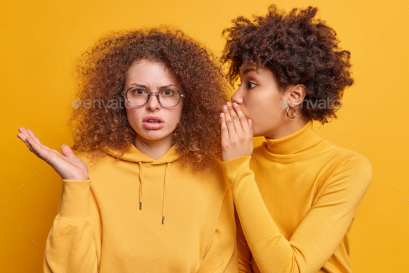 Surprised Afro American woman whispers secret to friend tells secretly something on ear share gossip