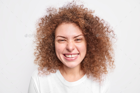 Close up shot of happy sincere European female model has fun smirks face from joy has curly bushy ha
