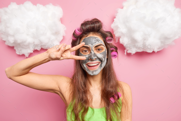 Positive brunette woman smiles gently makes peace gesture over eyes applies clay mask to reduce fine