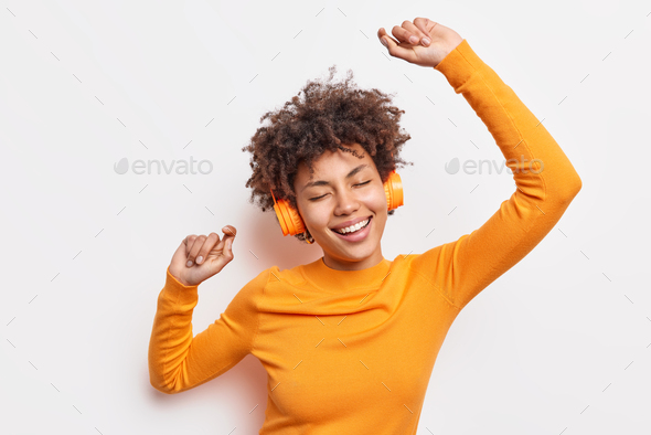 Happy young Afro American woman carried away with music dances carefree with arms raised keeps eyes