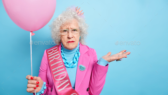 Studio shot of wrinkled grey haired elderly woman raises palms looks seriously at camera dressed in