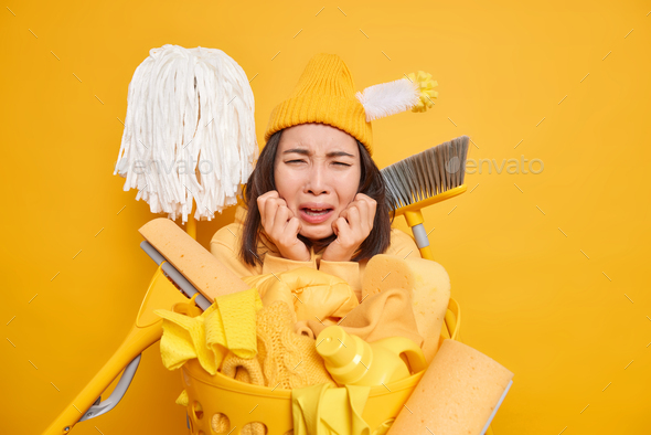 Dejected Asian woman being tired of tidying up house surrounded by cleaning equipment laundry basket