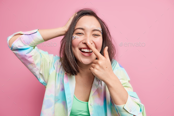 Young joyful Asian woman has fun smiles broadly touches nose expresses positive emotions has face de