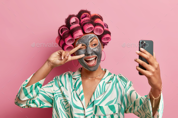 Photo of pleased ethnic woman makes peace gesture over eye smiles broadly applies beauty mask hair r