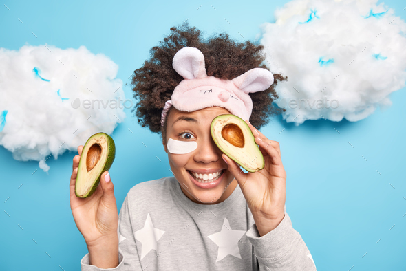 Photo of happy curly haired woman covers eyes with halves of avocado enjoys skin care procedures app