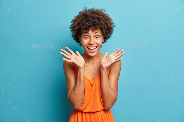 Positive dark skinned curly haired woman happy to receive good news spreads palms smiles broadly wea