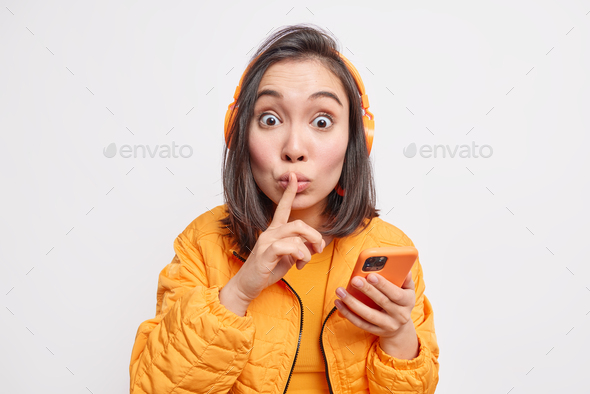 Studio shot of surprised woman gossips about something makes silence gesture tells secret holds mode