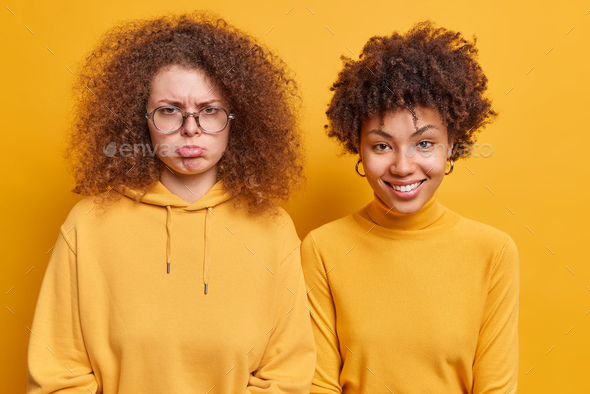 Two curly haired women stand next to each other express different emotions. Sad frustrated woman exp