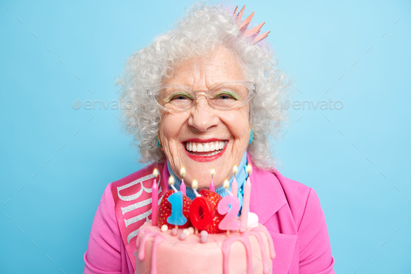 Horizontal shot of positive wrinkled European woman holds birthday cake dressed in stylish clothes f