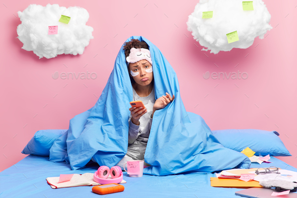 Dissatisfied sad woman wrapped in blanket waits for call holds smartphone wears pajama and beauty pa