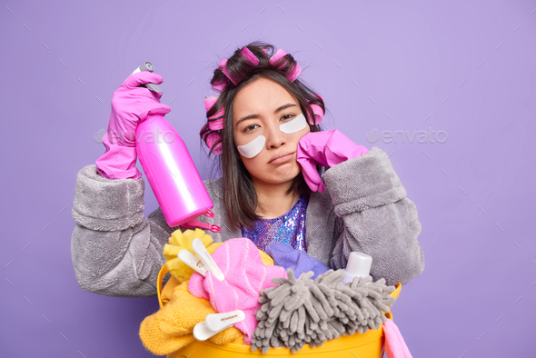 Tired Asian housemaid looks sadly at camera feels exhausted after cleaning house applies hair roller