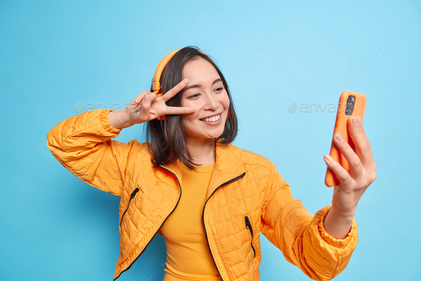 Happy Asian woman smiles gladfully makes peace gesture over eye takes selfie modern smartphone liste