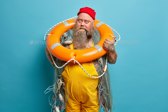 Stout experienced bearded fisherman smokes pipe poses with inflated ring and fishing nets indicates