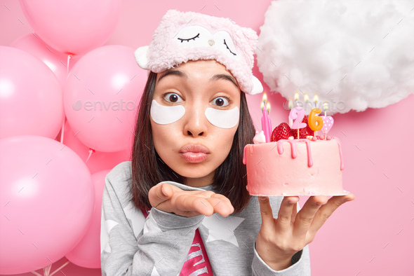Affectionate Asian woman blows air kiss at camera keeps lips folded holds festive cake with burning