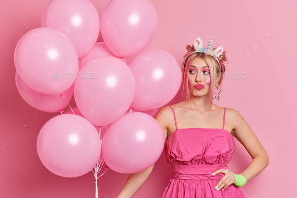Pensive woman with hairstyle dressed in festive dress holds bunch of balloons thinks about decoratio