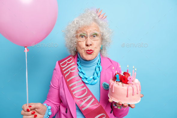 Pretty grey haired wrinkled woman keeps lips folded wears bright makeup holds delicious cake celebra