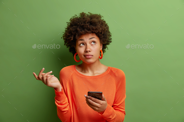 Perplexed hesitant curly haired African American woman shrugs shoulder being not sure uses smartphon