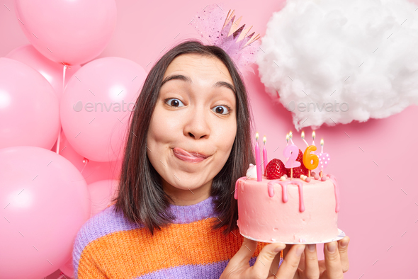 Close up shot of birthday girl holds tasty cake with candles licks lips celebrates 26th bday poses a