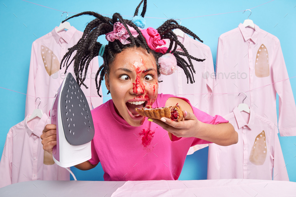 Funny crazy housewife bites delicious pie crosses eyes makes grimace has dreadlocks irons clothes at