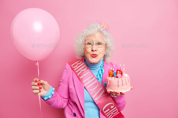 Aged curly haired senior wrinkled woman keeps lips rounded holds delicious cake with burning candles
