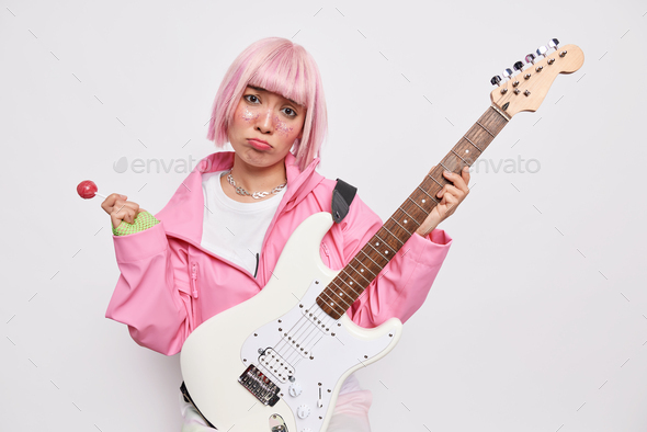 Upset teenage girl cannot learn playing guitar holds sweet lollipop bass acoustic guitar has pink ha