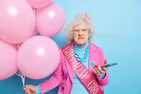 Dissatisfied wrinkled senior woman celebrates 100th birthday holds smartphone and bunch of inflated