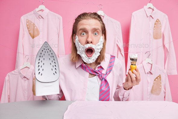 Amazed redhead adult man applies shaving gel on cheeks irons clothes dresses for work shocked to get
