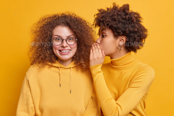 Two best friends gossip share secrets with each other. Afro American woman whispers gossips into com