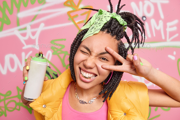Cheerful teen girl with dreadlocks golden teeth makes peace or victory gesture makes graffiti with a