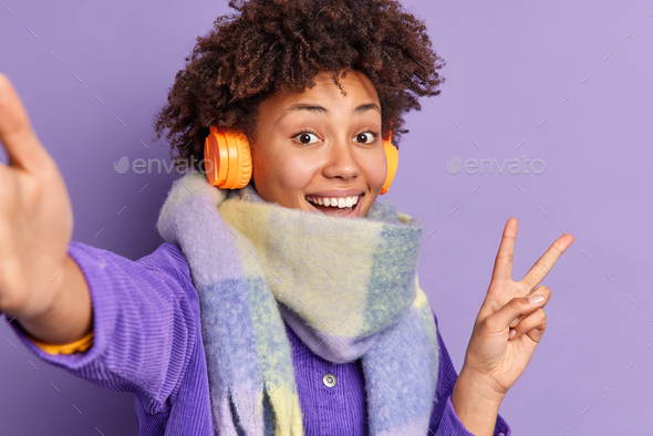 Positive Afro American woman smiles happily makes peace gesture while taking selfie dressed in jacke