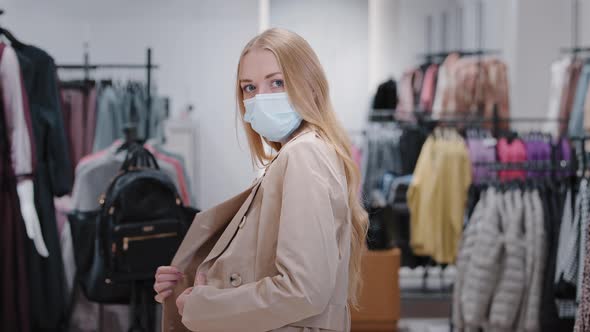 Millennial Woman in Face Medical Mask in Shopping Mall Chooses Autumn Jacket Coat New Outfit Trying alt