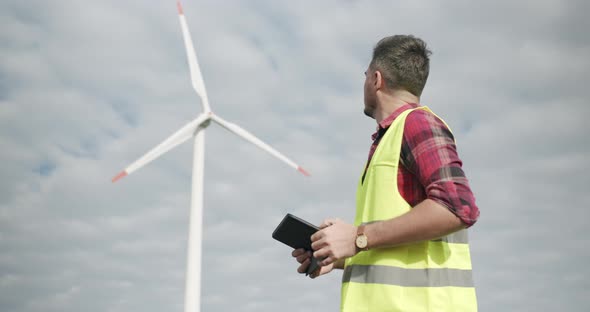 Experienced Engineer with Tablet Looks Behind on Wind Mill and Looking at Camera alt