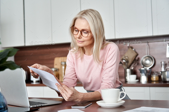 Middle aged woman housewife reading paper letter bill sitting at home kitchen.
