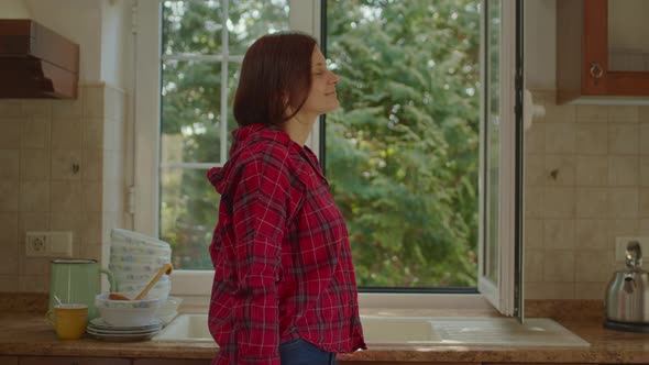 Young Adult Woman Enjoying Fresh Summer Say Standing By the Opened Window in the Kitchen alt
