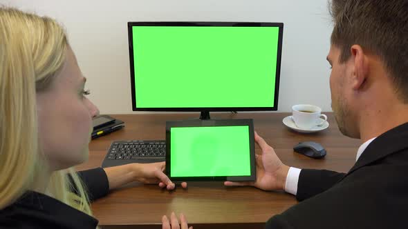 Two office workers sit at a desk and look at a computer and a tablet with green screens  alt