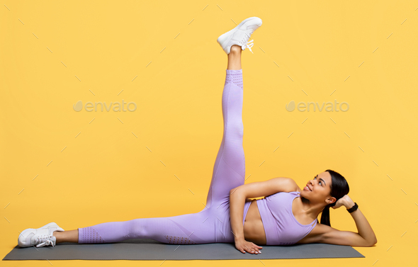 Fitness african american woman stretching and lifting leg up, exercising on mat over yellow