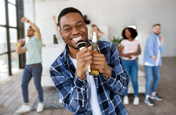 Happy black guy holding bottle of beer and microphone, performing song, singing karaoke on party
